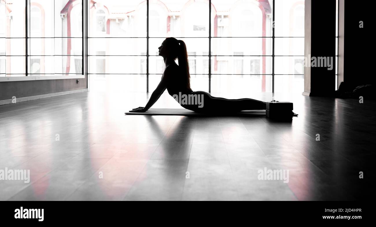 Junge lateinerin praktiziert im Yoga-Studio, Silhouette. Basic Yoga Cobra Pose liegend auf einer Sportmatte. Fitness und Yoga Stockfoto