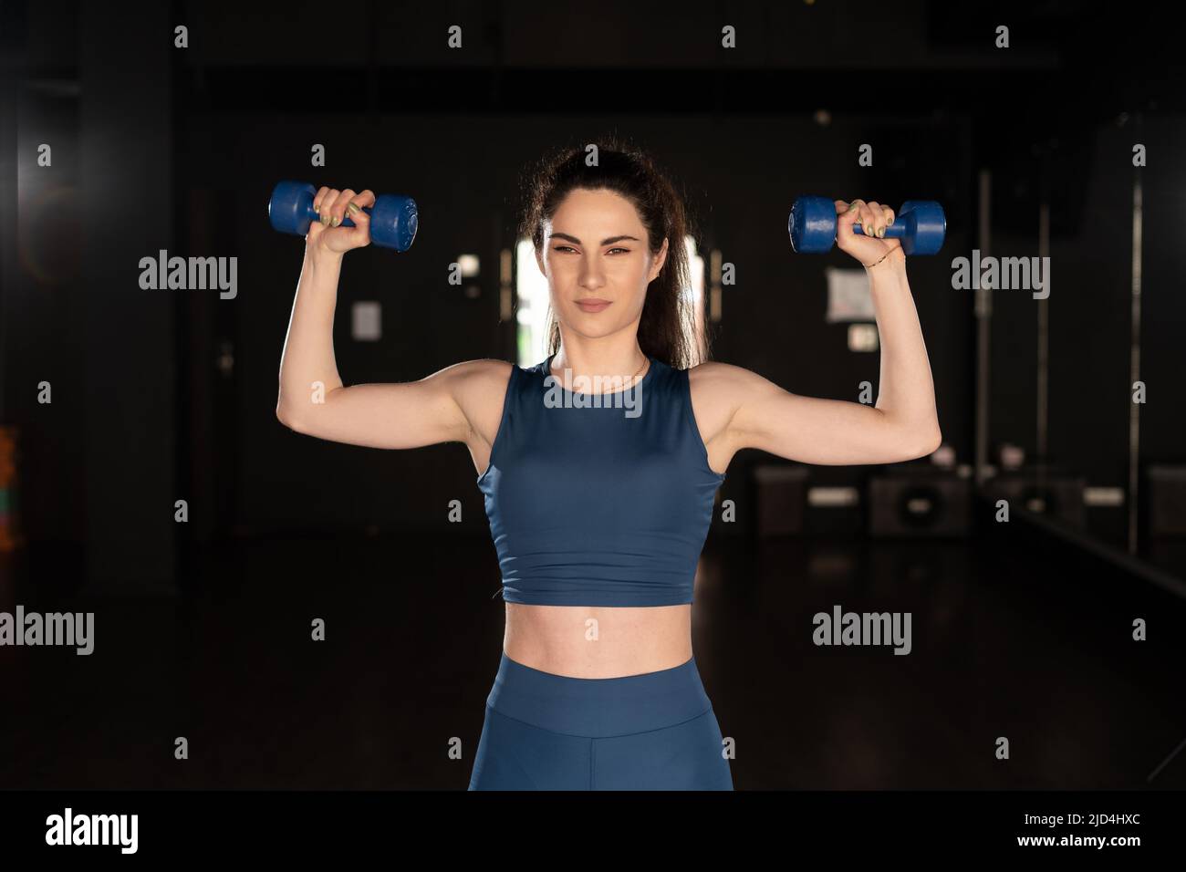 Entschlossene Frau, die im Fitnessstudio Gewicht verliert und mit Kurzhanteln trainiert. Schöne latinerin in Sportkleidung mit blauen Hanteln in den Händen. Schauen Sie zu Stockfoto