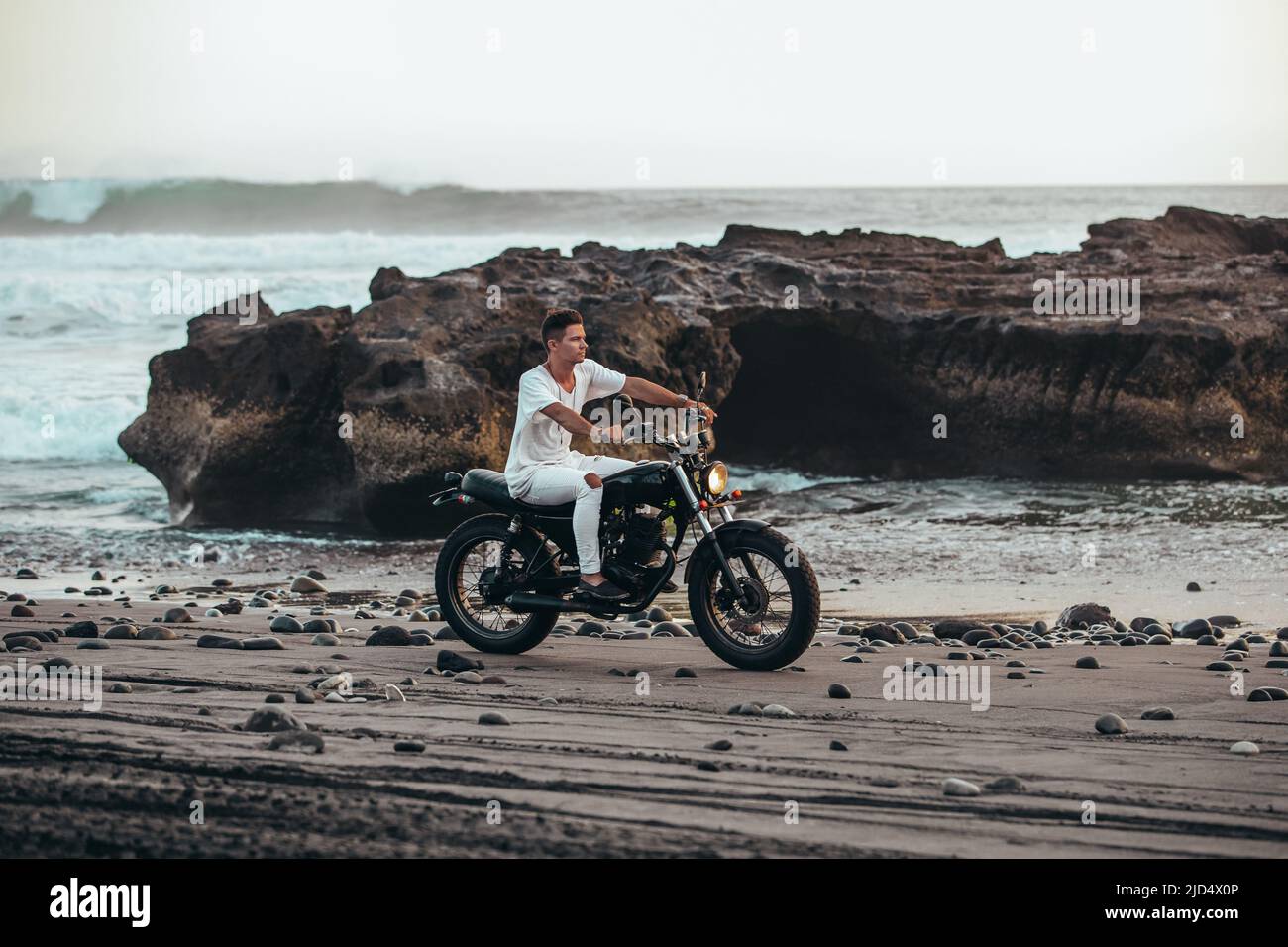 Junger Mann in weißem Outfit, der während des Sonnenuntergangs auf dem schwarzen Sandstrand von Bali auf einem Vintage-Motorrad reitet Stockfoto