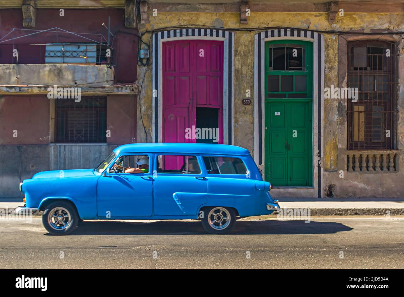 Ein hellblauer Oldtimer fährt an einer violetten und einer grünen Haustüre in der Altstadt von Havanna, Kuba vorbei Stockfoto