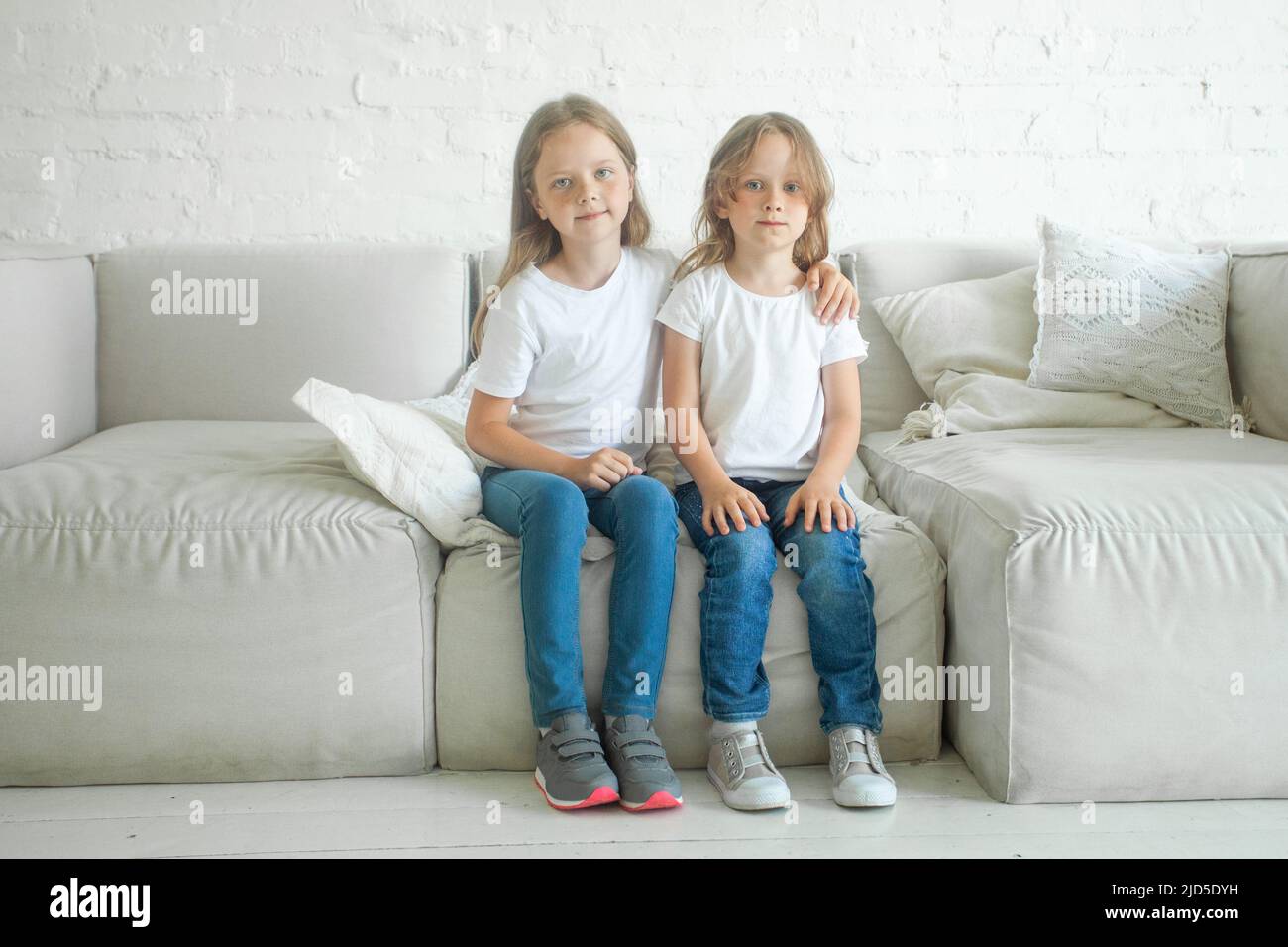 Zwei kleine Mädchen Schwestern umarmen und sitzen auf dem Sofa drinnen Stockfoto
