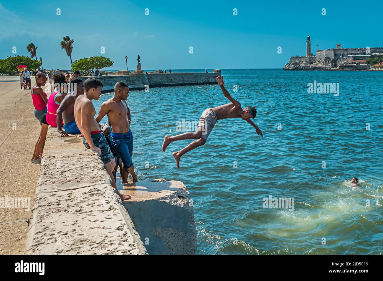 Jugendliche springen von einer Mauer ins Wasser auf dem Malecon in Havanna, Kuba mit der berühmten Festung El Morro im Hintergrund Stockfoto