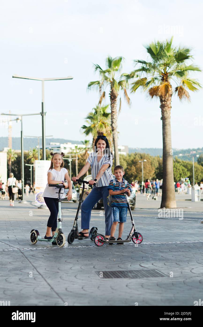 Fröhlicher Familienleben und Urlaubskonzept. Mutter, kleiner Junge, Mädchen, Roller reiten, in der Altstadt spazieren gehen, Straße. Lachen an einem sonnigen Sommertag. Stockfoto