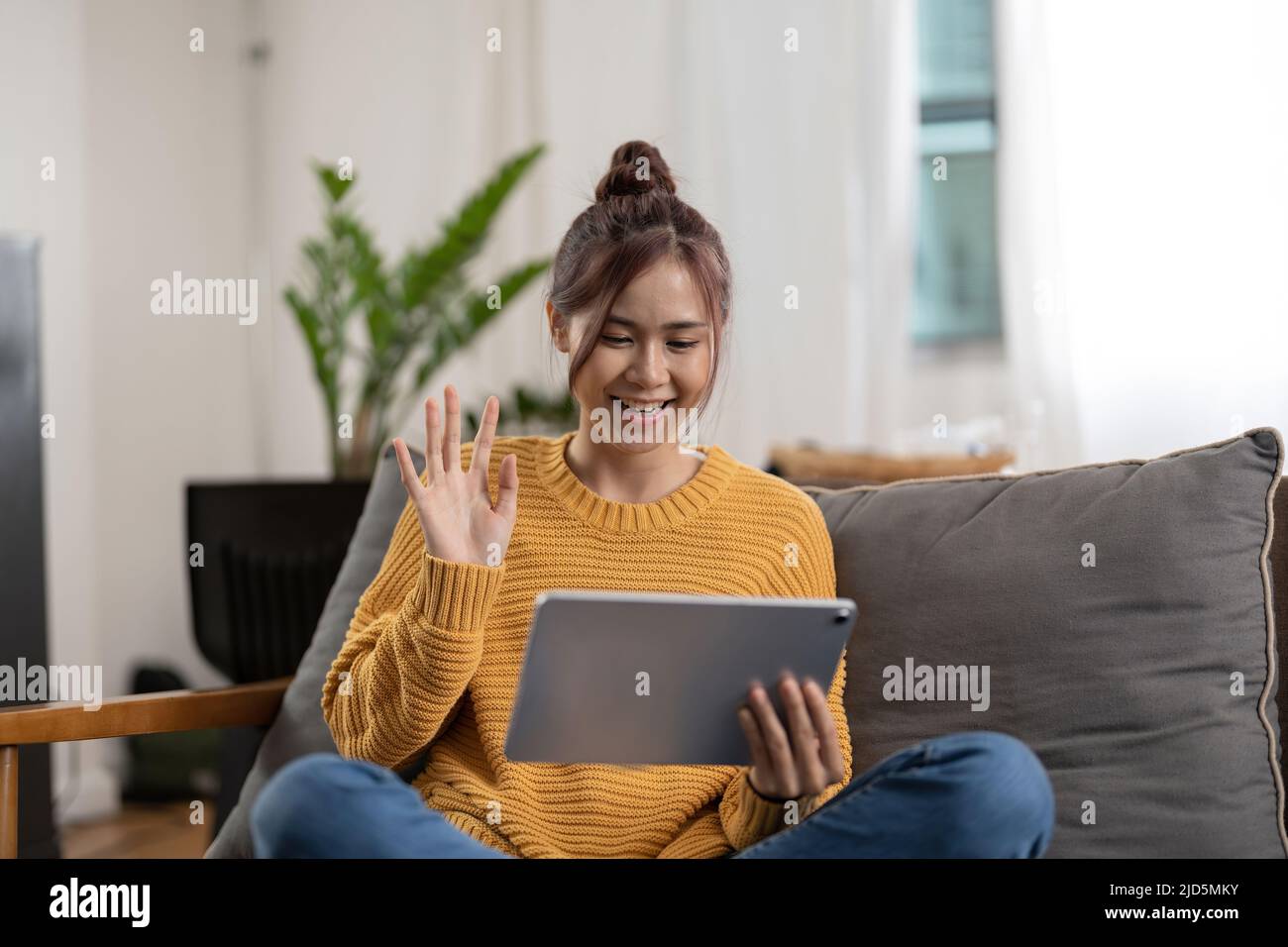 Glücklich lächelnde asiatische junge Frau in gelben Pullover mit Tablet-pc-Computer mit Videoanruf und winken Hand zu Hause Stockfoto