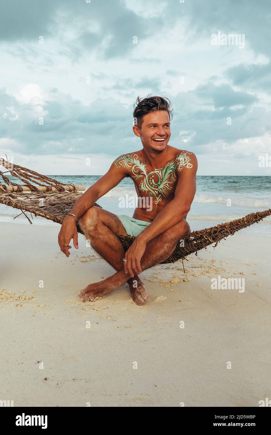 Junger, fröhlich lächelnder Mann mit türkisfarbener Körperfarbe, der während des Sonnenuntergangs auf einer Seilhängematte am Strand sitzt Stockfoto