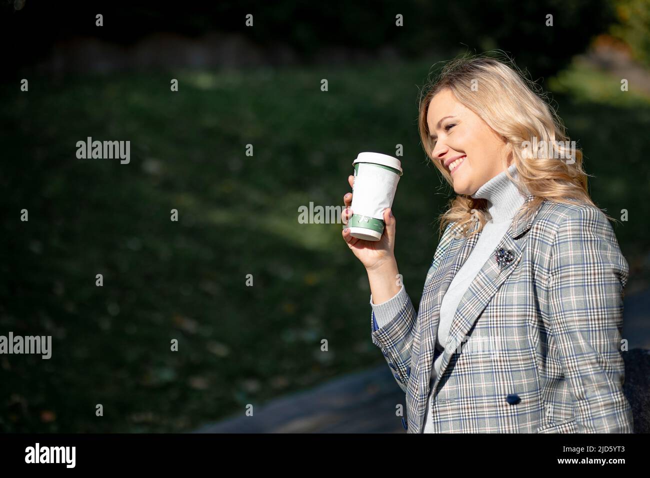 Charmante Blondine mit langen lockigen Haaren in grau-blauer Tweed-Jacke und passendem Kaschmirpullover, die eine Tasse Tee in den Händen hält, während sie bei Sonnenschein geht Stockfoto