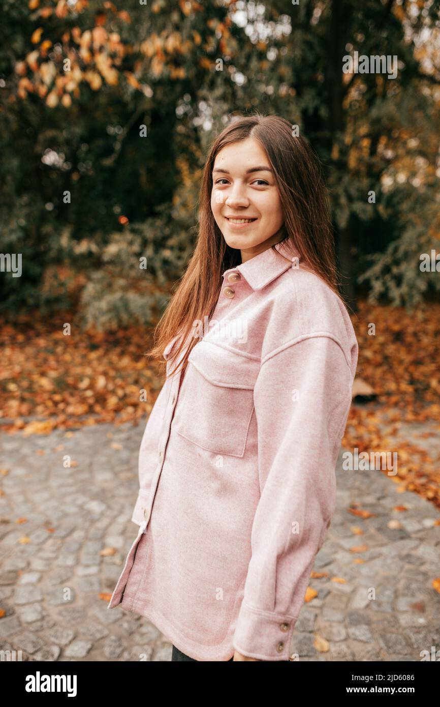 Charmante, braunhaarige Frau, die an einem warmen Herbsttag auf dem Stadtplatz spazieren geht, sieht glücklich und friedlich aus. Sie trägt ein warmes, weiches rosafarbenes Hemd. Wochenende Stockfoto