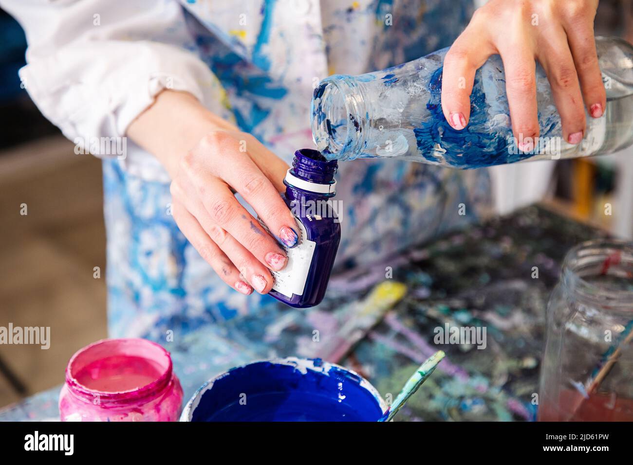 Beschnittenes Foto von Händen einer Frau in einem Gewand, bedeckt mit Flecken, die Wasser aus dem Glas in eine kleine Plastikflasche gießen. Stockfoto