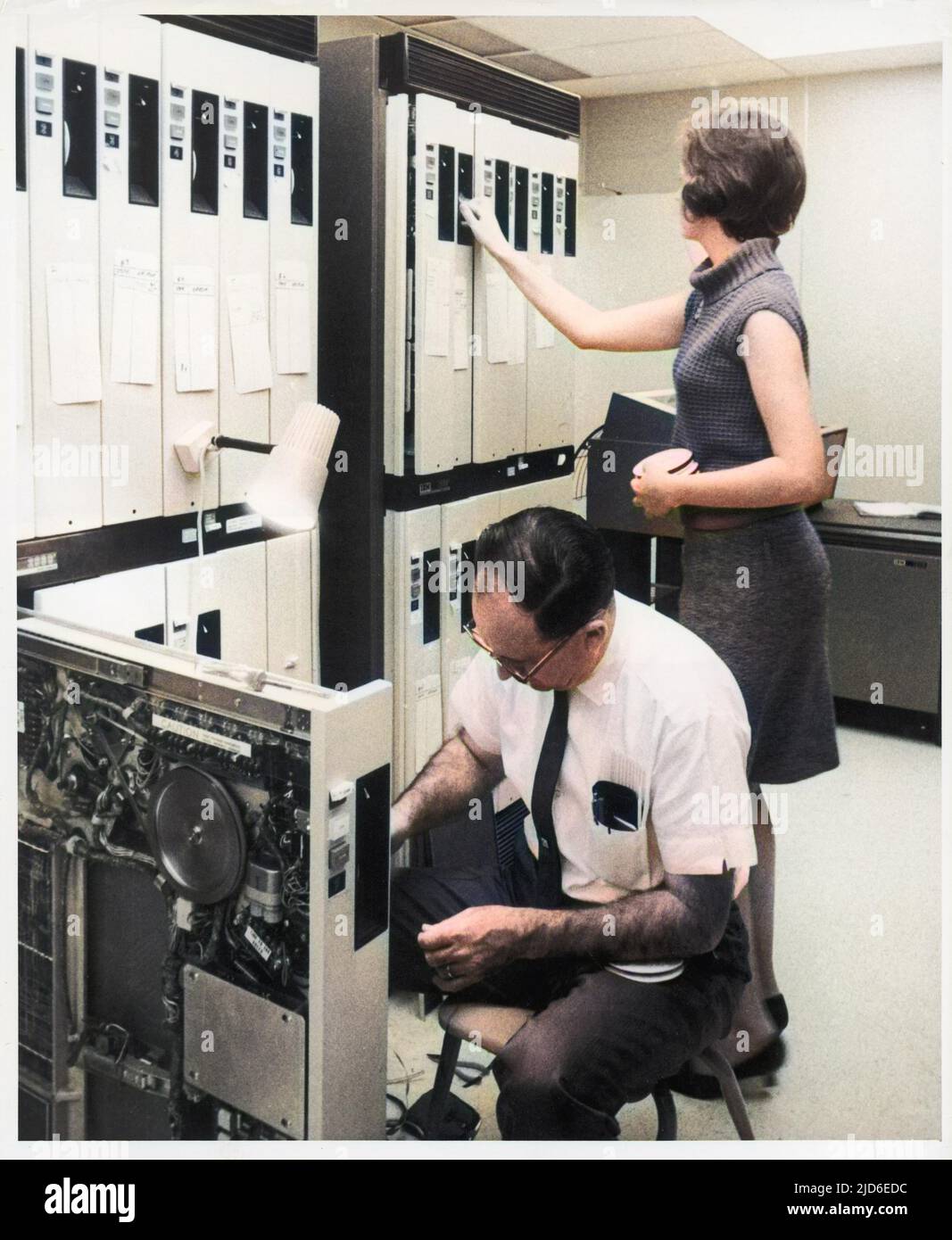Ein männlicher Trouble-Shooter entwirrt in einem Terminal ein eingeklemmtes Band, während eine Lehrerin in einem anderen Terminal im riesigen Computerraum einer Schule oder Hochschule die Rollen wechselt. Kolorierte Version von : 10144583 Datum: 1960s Stockfoto