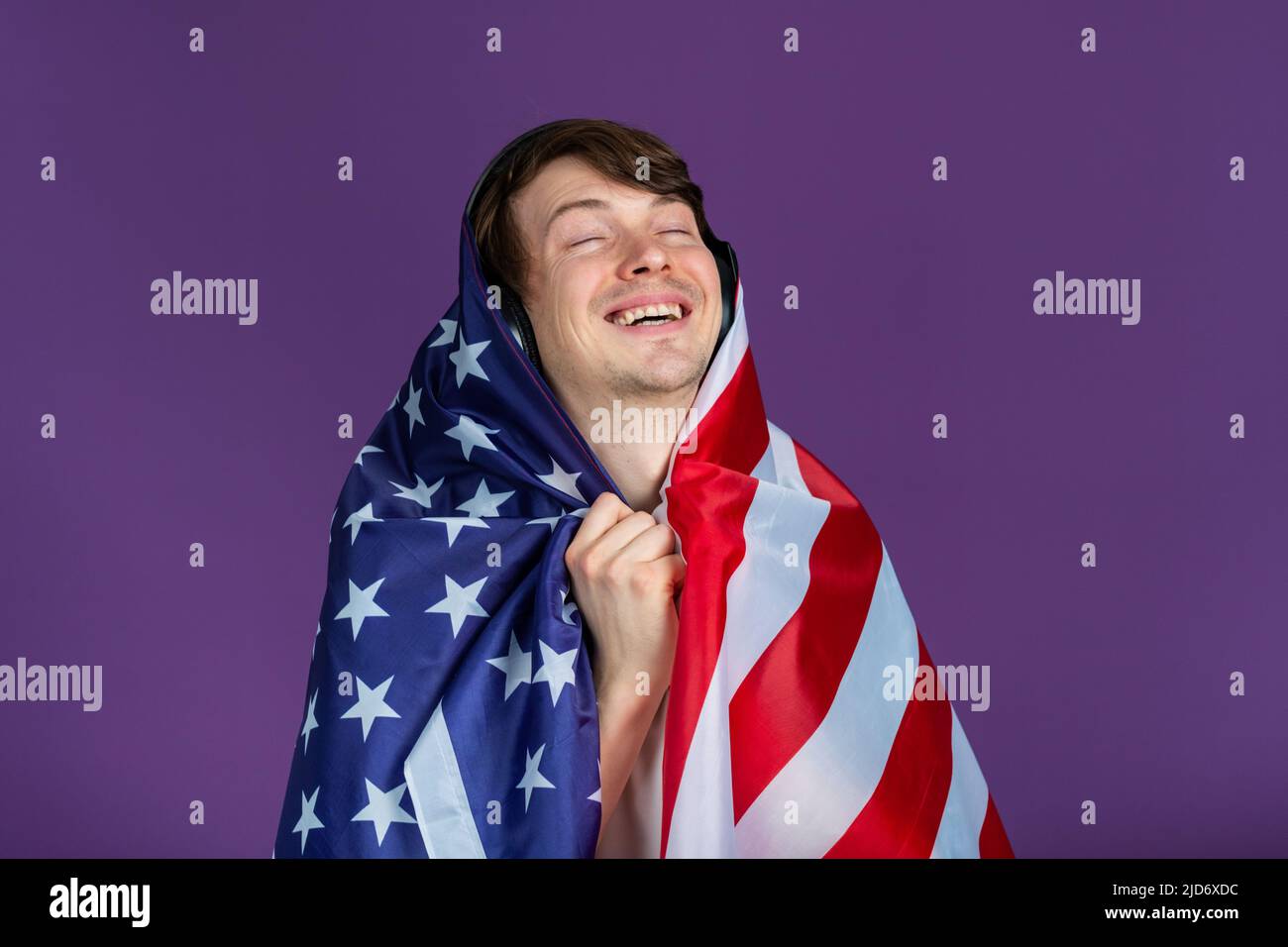 Portrait eines glücklichen Mannes mit Kopfhörern, der in eine amerikanische Flagge verwandelt wurde, auf einem violetten Hintergrund, der Musik hört und genießt Stockfoto