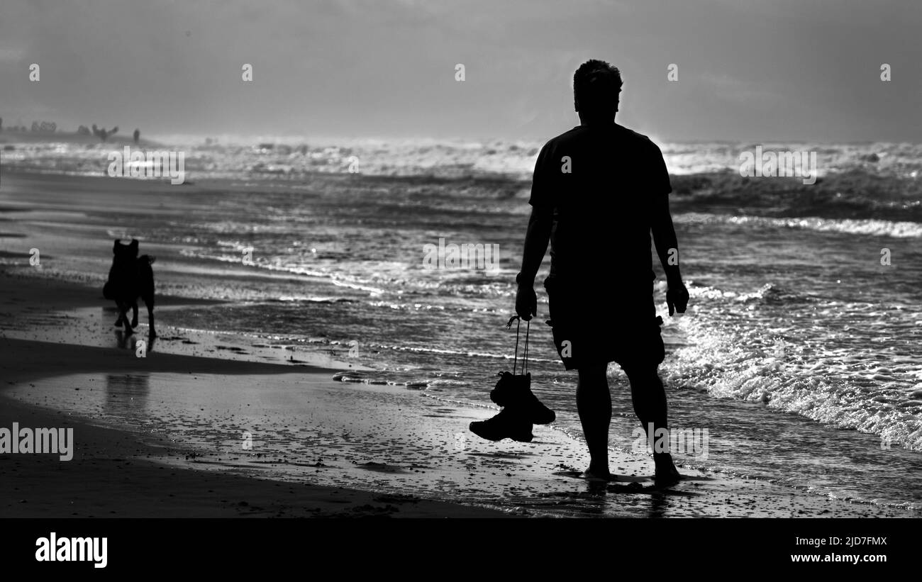 Schwarz-weißes Foto eines Mannes und seines Hundes, der am Ufer des Meeres steht und seine Schuhe in der Hand hält. Stockfoto