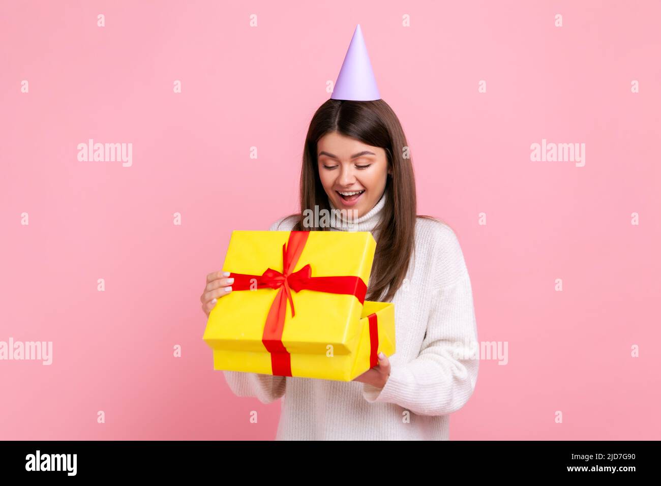 Positive glückliche Mädchen in Partei-Kegel Blick in eingewickelten Geschenkbox, offenes Geschenk, angenehme Überraschung, trägt weißen lässigen Stil Pullover. Innenaufnahme des Studios isoliert auf rosa Hintergrund. Stockfoto