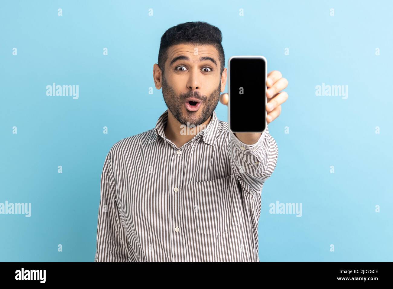 Staunender bärtiger Geschäftsmann, der sein Handy hochhielt und mit offenem Mund die Kamera anlächelte, das Gadget empfahl und ein gestreiftes Hemd trug. Innenaufnahme des Studios isoliert auf blauem Hintergrund. Stockfoto