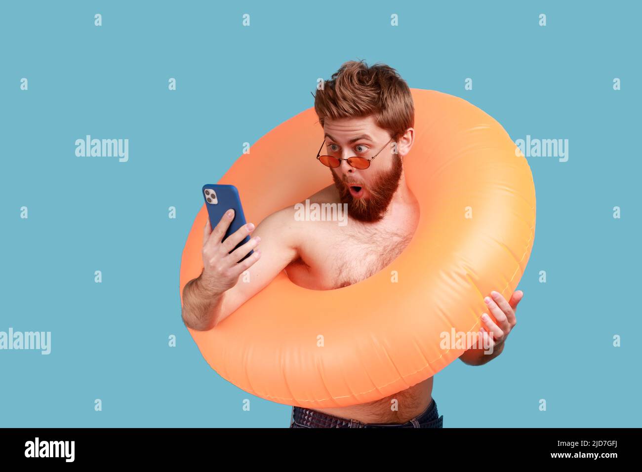 Porträt eines erstaunt überraschten bärtigen Mannes in Sonnenbrille, der mit orangefarbenem Gummiring steht und das Mobiltelefon hält und mit offenem Mund auf das Display blickt. Innenaufnahme des Studios isoliert auf blauem Hintergrund. Stockfoto