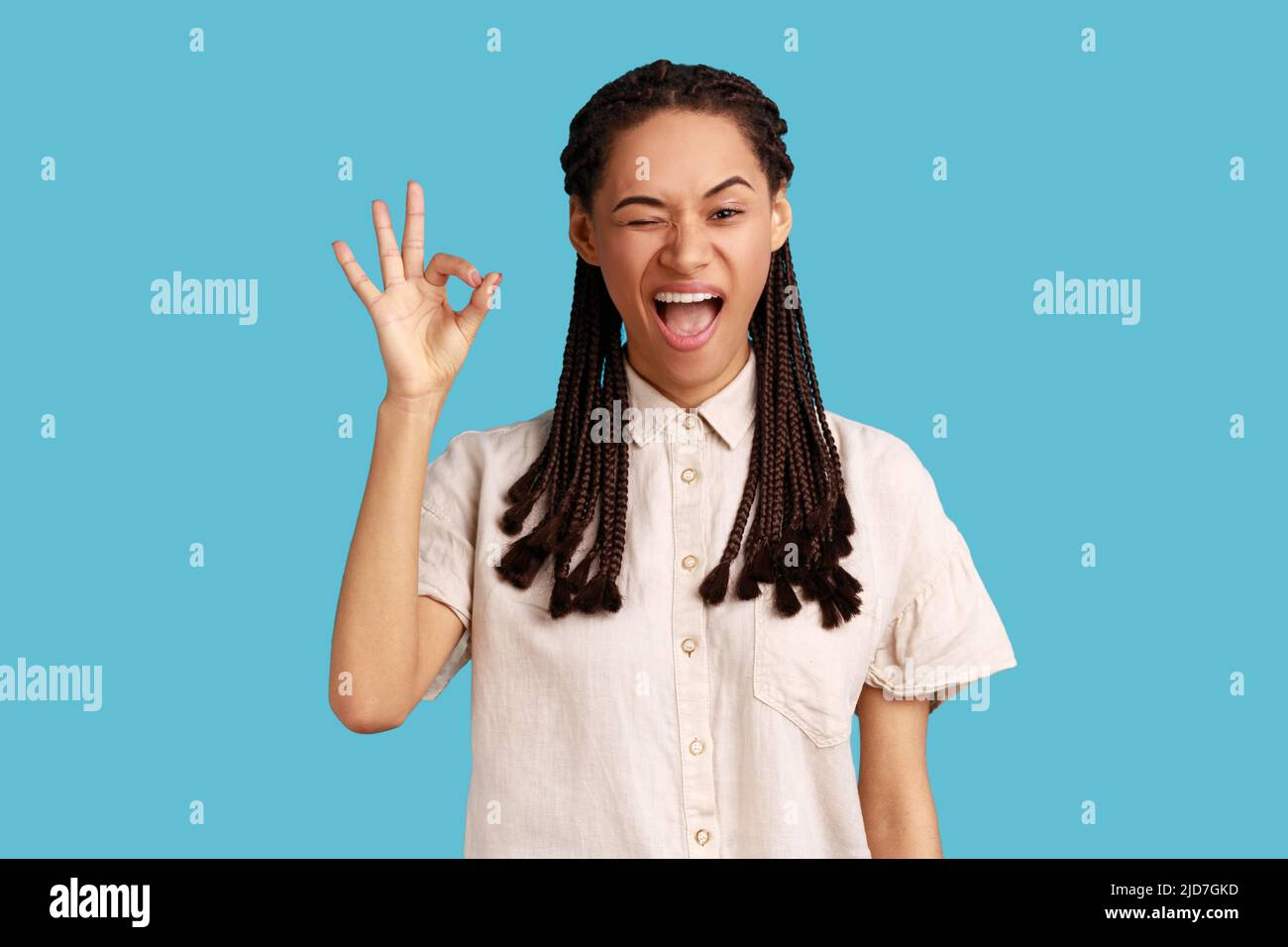 Portrait einer fröhlichen jungen erwachsenen Frau mit schwarzen Dreadlocks, die ein gutes Zeichen zeigt, versichert, dass alles in Ordnung ist, sieht gerne aus, trägt weißes Hemd. Innenaufnahme des Studios isoliert auf blauem Hintergrund. Stockfoto