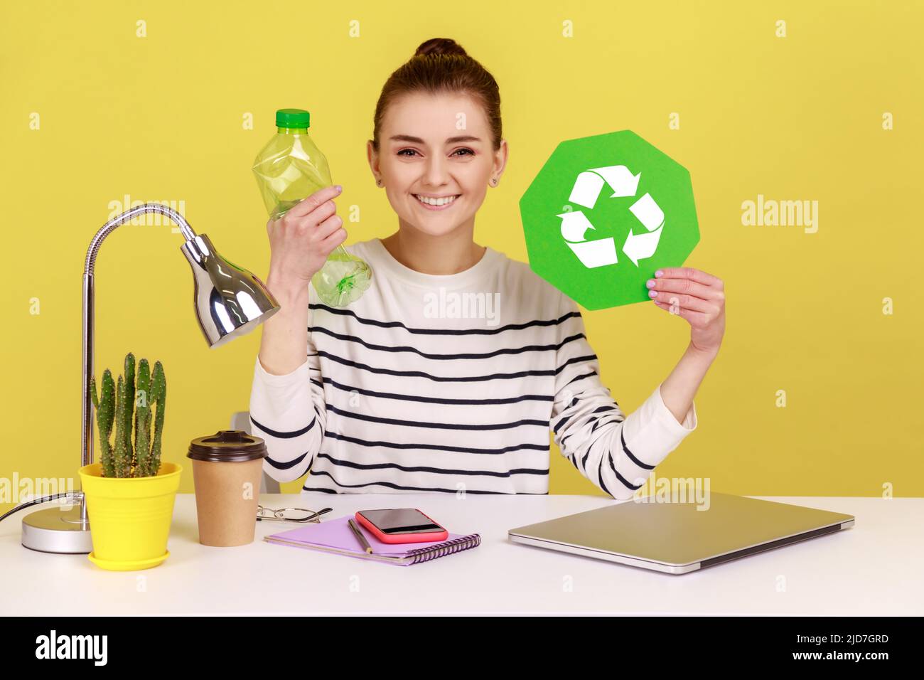 Porträt einer zufriedenen Frau mit einem charmanten Lächeln, die leere Plastikflasche und grünes Recycling-Schild am Arbeitsplatz hält. Studio-Innenaufnahme isoliert auf gelbem Hintergrund. Stockfoto