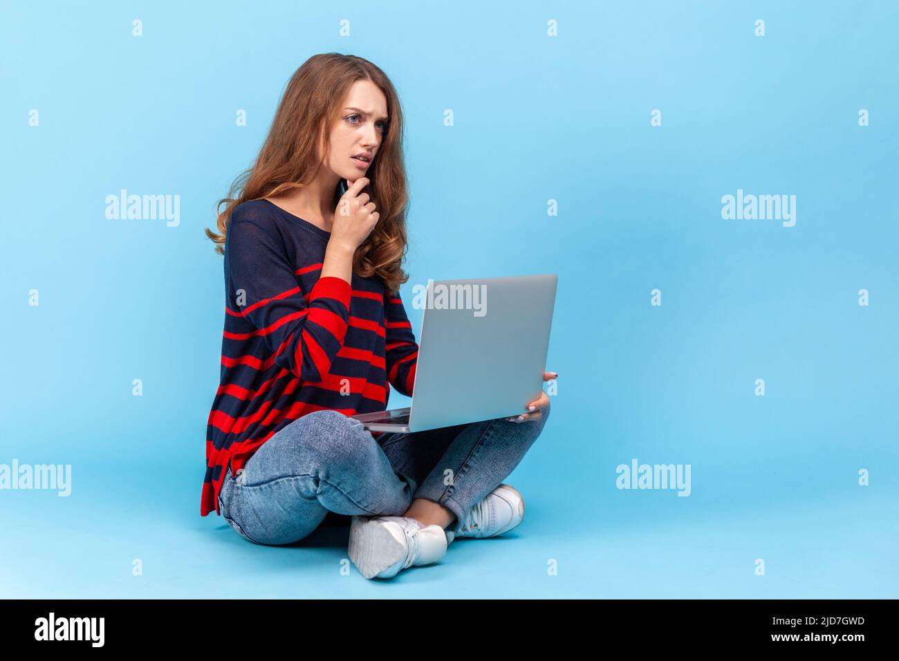 Nachdenkliche Frau mit gestreiftem Pullover im lässigen Stil, sitzend mit gekreuzten Beinen, Laptop auf den Knien haltend, online arbeitend, nachdenklich ausdruckend. Innenaufnahme des Studios isoliert auf blauem Hintergrund. Stockfoto