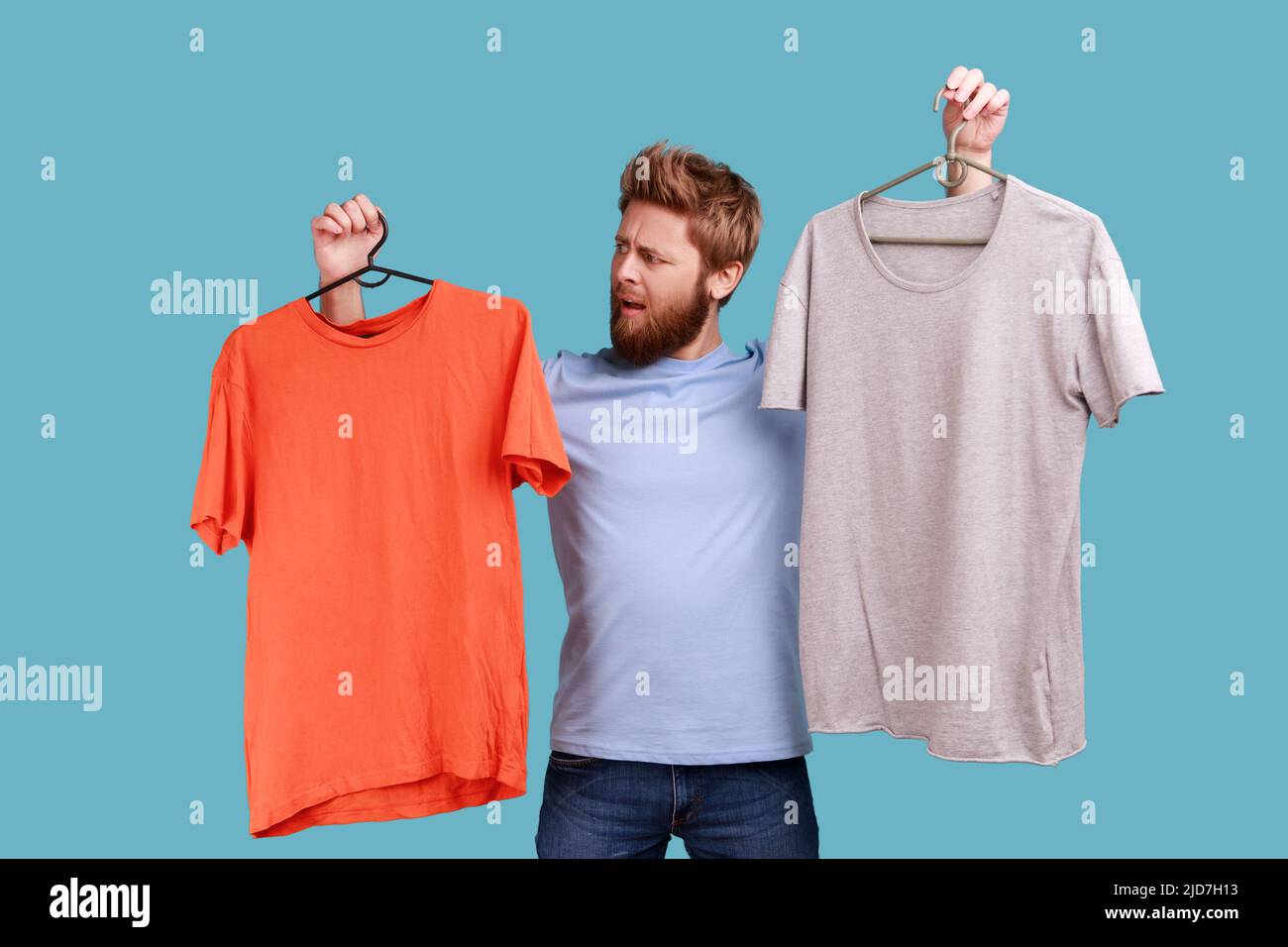 Porträt eines verwirrten bärtigen Mannes mit runzelndem Gesicht, der zwei Kleiderbügel mit grauen und orangefarbenen T-Shirts hält, die schwer zu wählen sind, um die beste Kleidung zu finden. Innenaufnahme des Studios isoliert auf blauem Hintergrund. Stockfoto