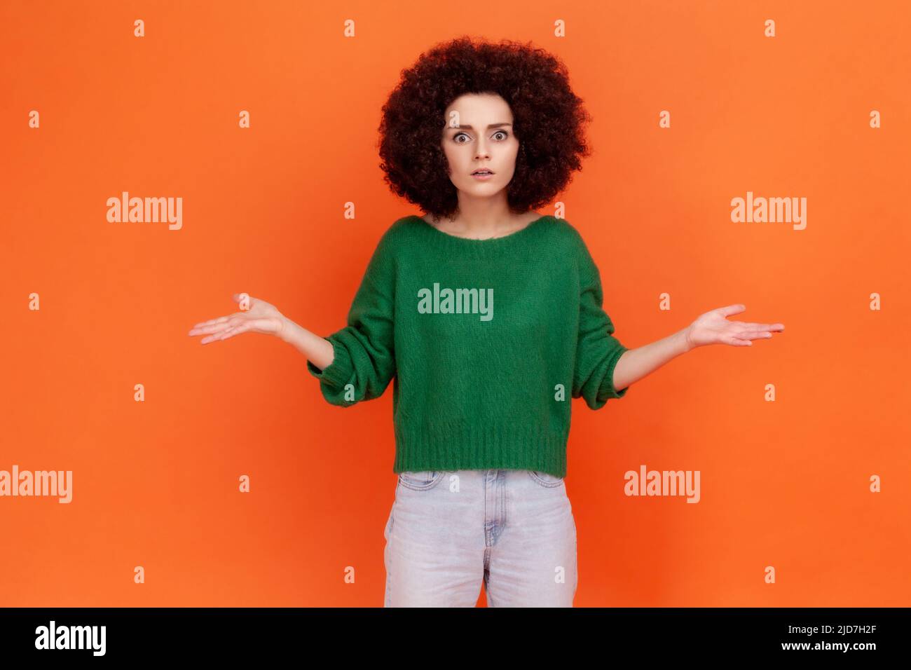 Wütende Frau mit Afro-Frisur trägt einen grünen Pullover im Casual-Stil, der mit erhobenen Händen und empörendem Ausdruck steht und den Grund des Konflikts fragt. Innenaufnahme des Studios isoliert auf orangefarbenem Hintergrund. Stockfoto