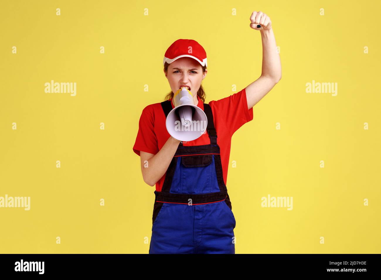 Porträt einer aggressiven Arbeiterin, die mit wütender Mimik etwas schreit, protestiert, die Faust hochhebt, Overalls und rote Mütze trägt. Innenaufnahme des Studios isoliert auf gelbem Hintergrund. Stockfoto