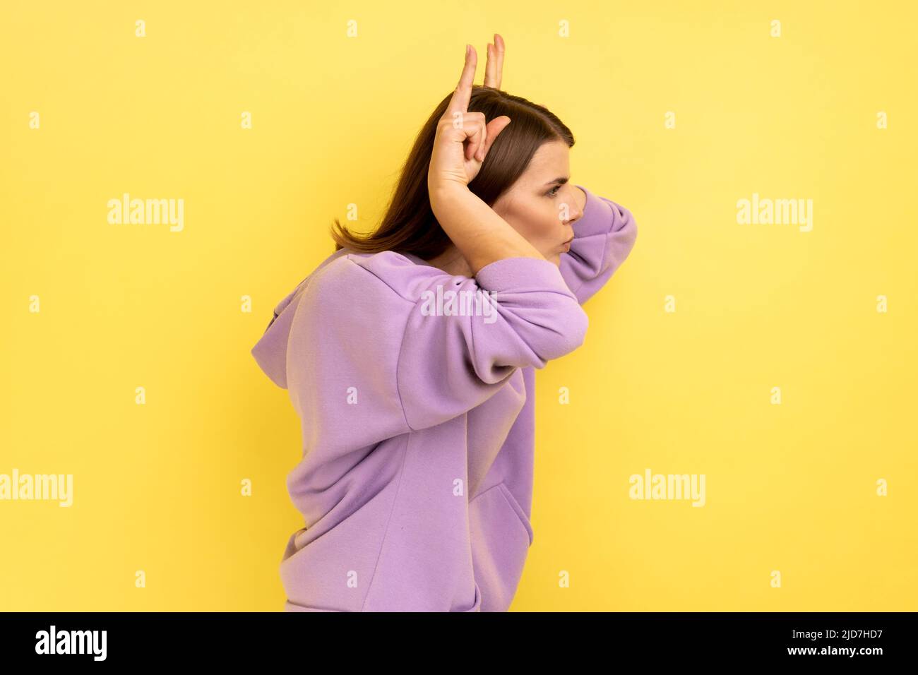 Seitenansicht einer aggressiven Frau, die die Finger über den Kopf hält und Hörner zeigt, arrogant und stur, angriffbereit, mit violettem Hoodie. Innenaufnahme des Studios isoliert auf gelbem Hintergrund. Stockfoto