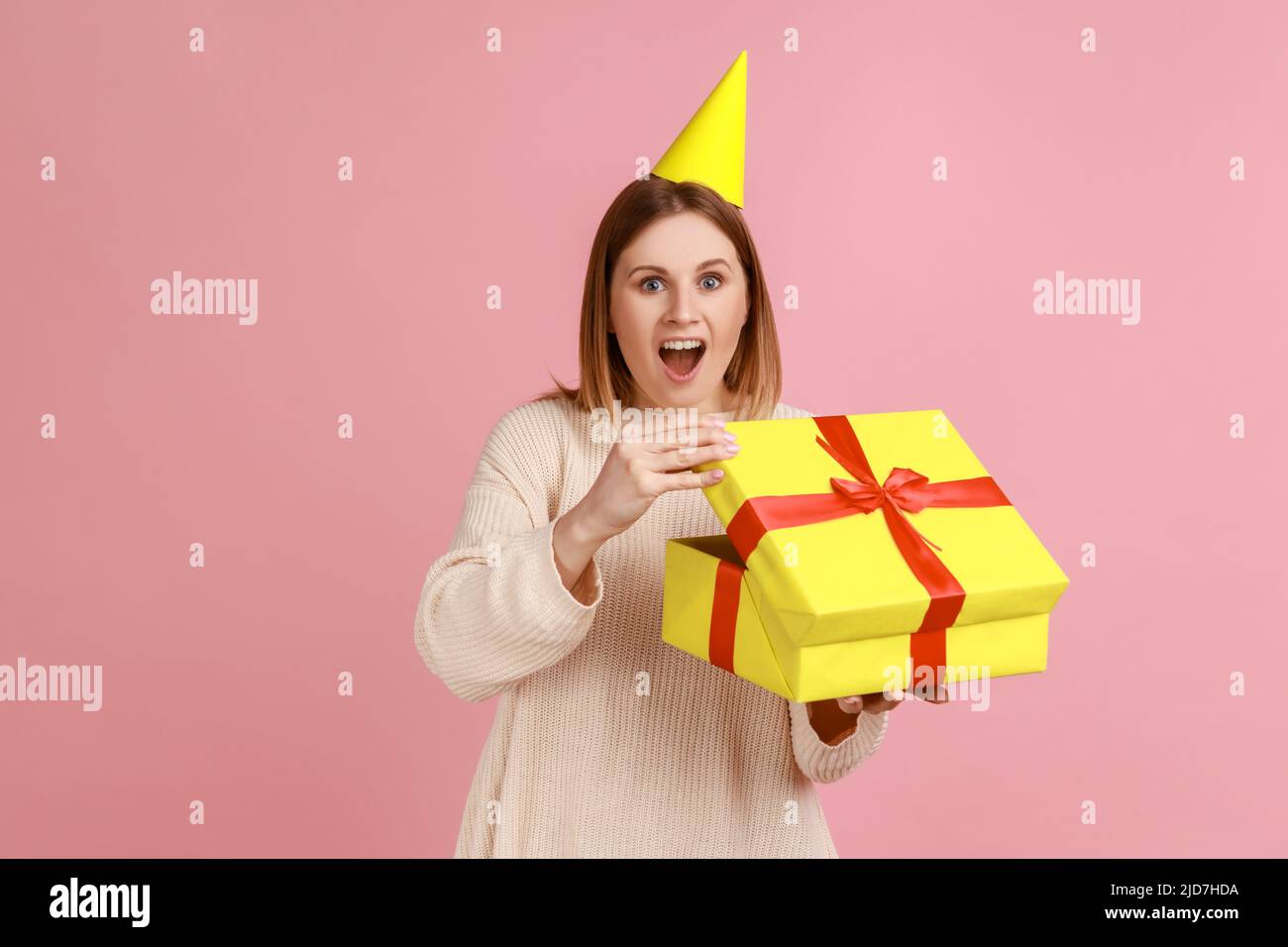 Porträt der aufgeregt erstaunt blonde Frau in Partei-Kegel Auspacken Geschenk-Box, mit angenehmer Überraschung, Blick auf die Kamera, trägt weißen Pullover. Innenaufnahme des Studios isoliert auf rosa Hintergrund. Stockfoto