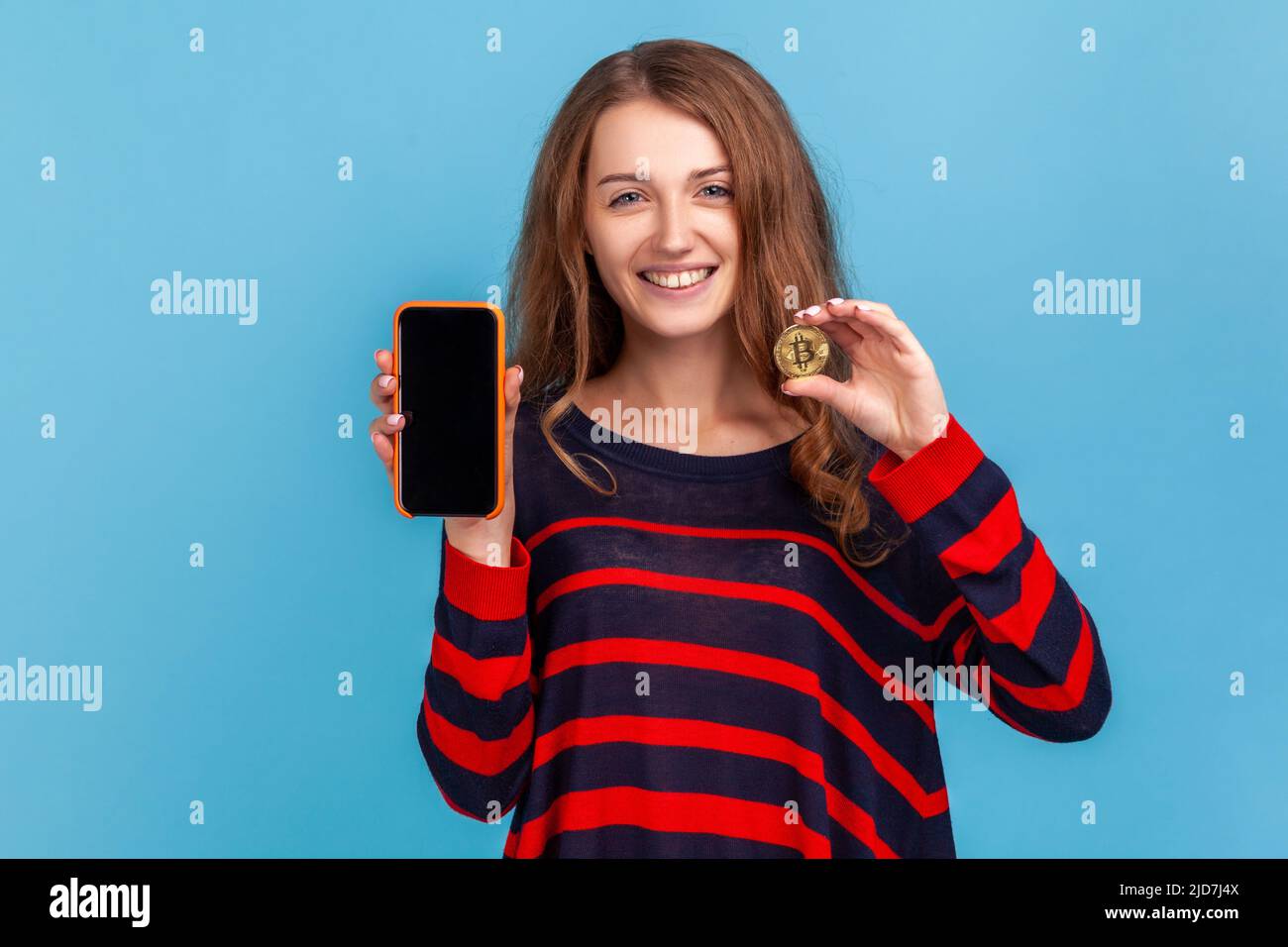 Fröhliche Frau in gestreiftem Pullover im lässigen Stil, mit goldenem Bitcoin und Smartphone mit leerem Bildschirm für Werbung, lächelnd vor der Kamera. Innenaufnahme des Studios isoliert auf blauem Hintergrund. Stockfoto