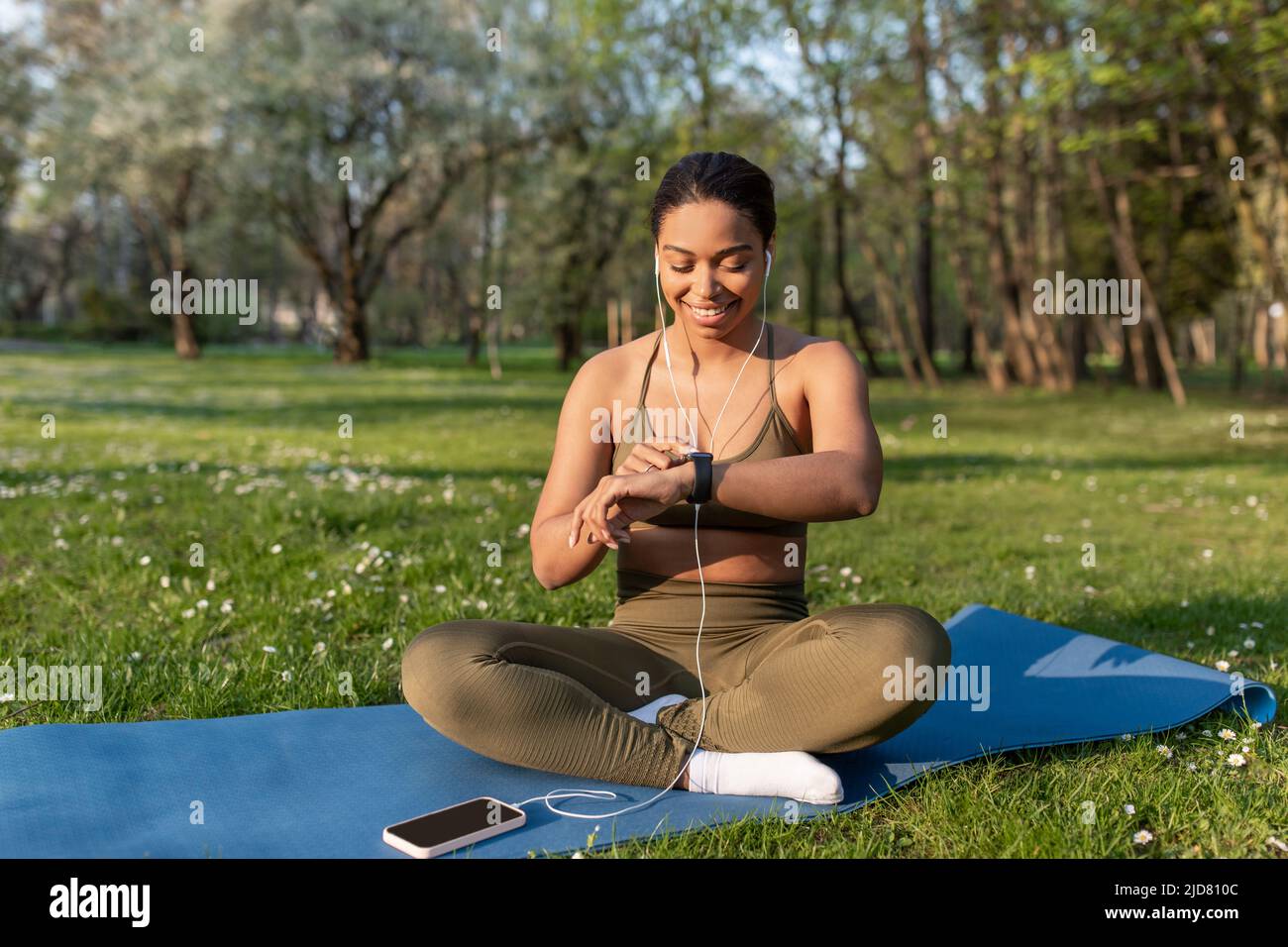 Junge schwarze Frau, die Mobiltelefon benutzt, Musik hört, Fitness Tracker während des Yoga-Trainings im Park überprüft Stockfoto