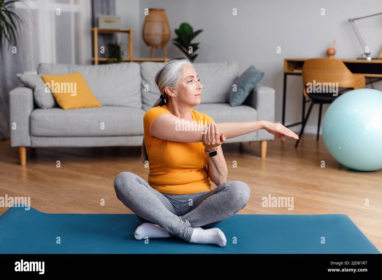 Aktive ältere Frau, die ihren Arm zu Hause auf einer Matte streckt, Flexibilitätsübungen macht, Yoga praktiziert oder Pilates praktiziert Stockfoto