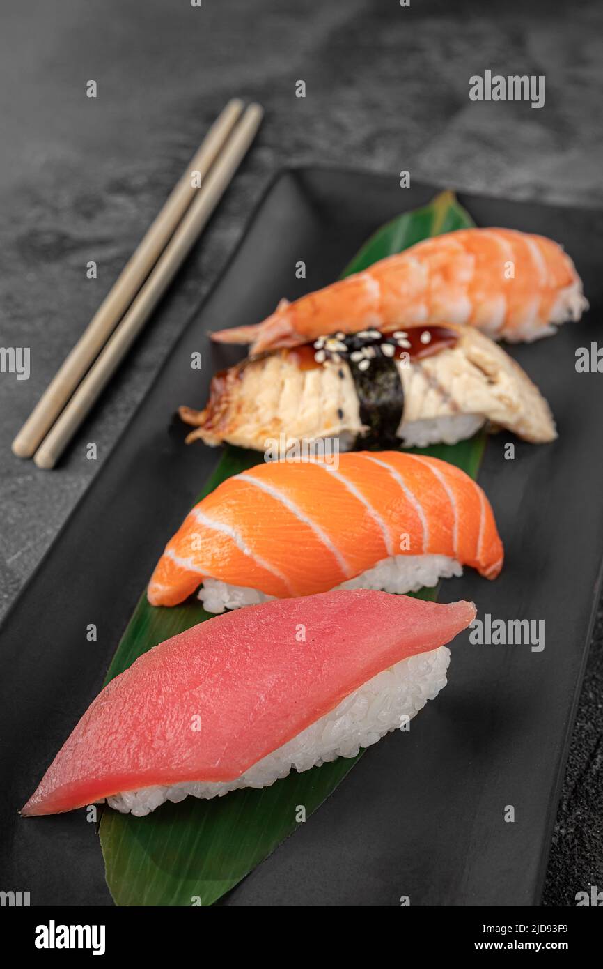 Set aus Nigiri mit Tiger Shrimp, tomago, Lachs, Thunfisch und grünem Bambusblatt in einer schwarzen Keramikplatte mit Essstäbchen auf einem dunkelgrauen strukturierten Hintergrund Stockfoto