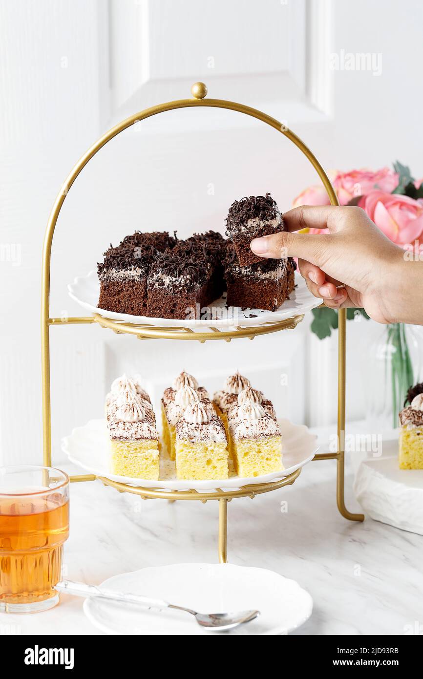 Weibliche Hand nehmen Schokoladenkuchen aus einem High Tea Rier Kuchen, Konzept English Afternoon Tea auf weißem Hintergrund Stockfoto