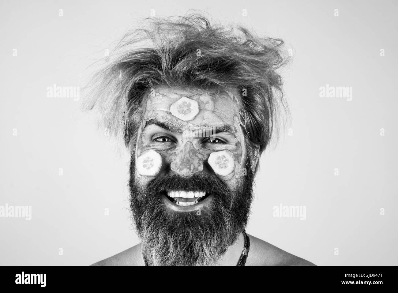 Happy man mit lustigen Gesichtsmaske mit Gurke Spaß haben. Verfahren zum Auftragen der Maske aus Ton auf das Gesicht des lustigen Mannes. Spa im Haus. Stockfoto