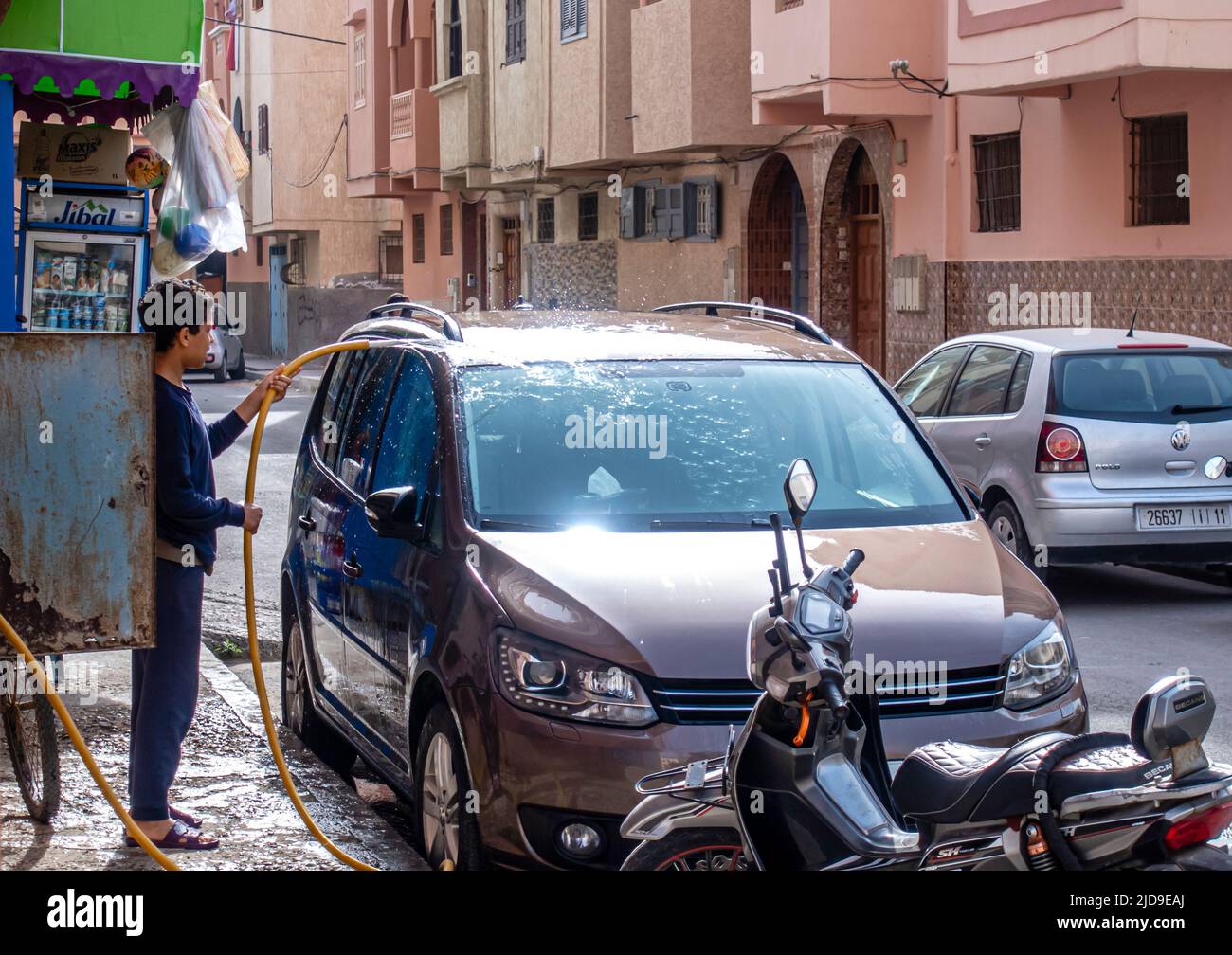 Ein Kind, das ein Auto mit Wasser aus einem Schlauch wäscht. Kinder, die in den Straßen von Essaouira, Marokko, arbeiten Stockfoto