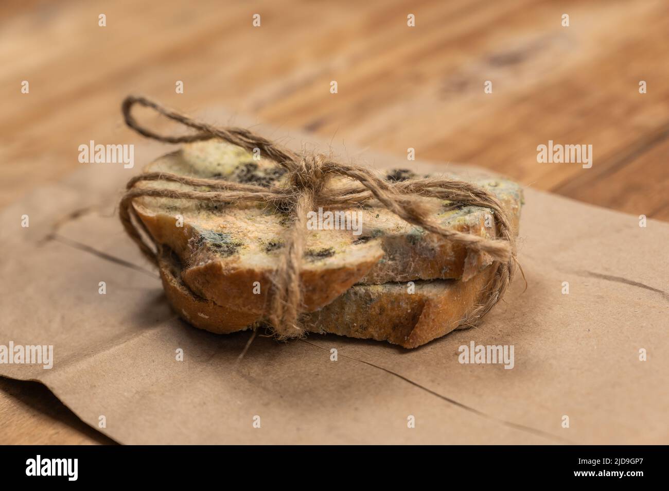 Schimmeliges Brot vor einem hölzernen Hintergrund. Zwei Scheiben Weizenbrot mit Pilzschimmel bedeckt. Die Scheiben sind mit Garn gebunden. Brauner Papierbeutel. Verdorben für Stockfoto