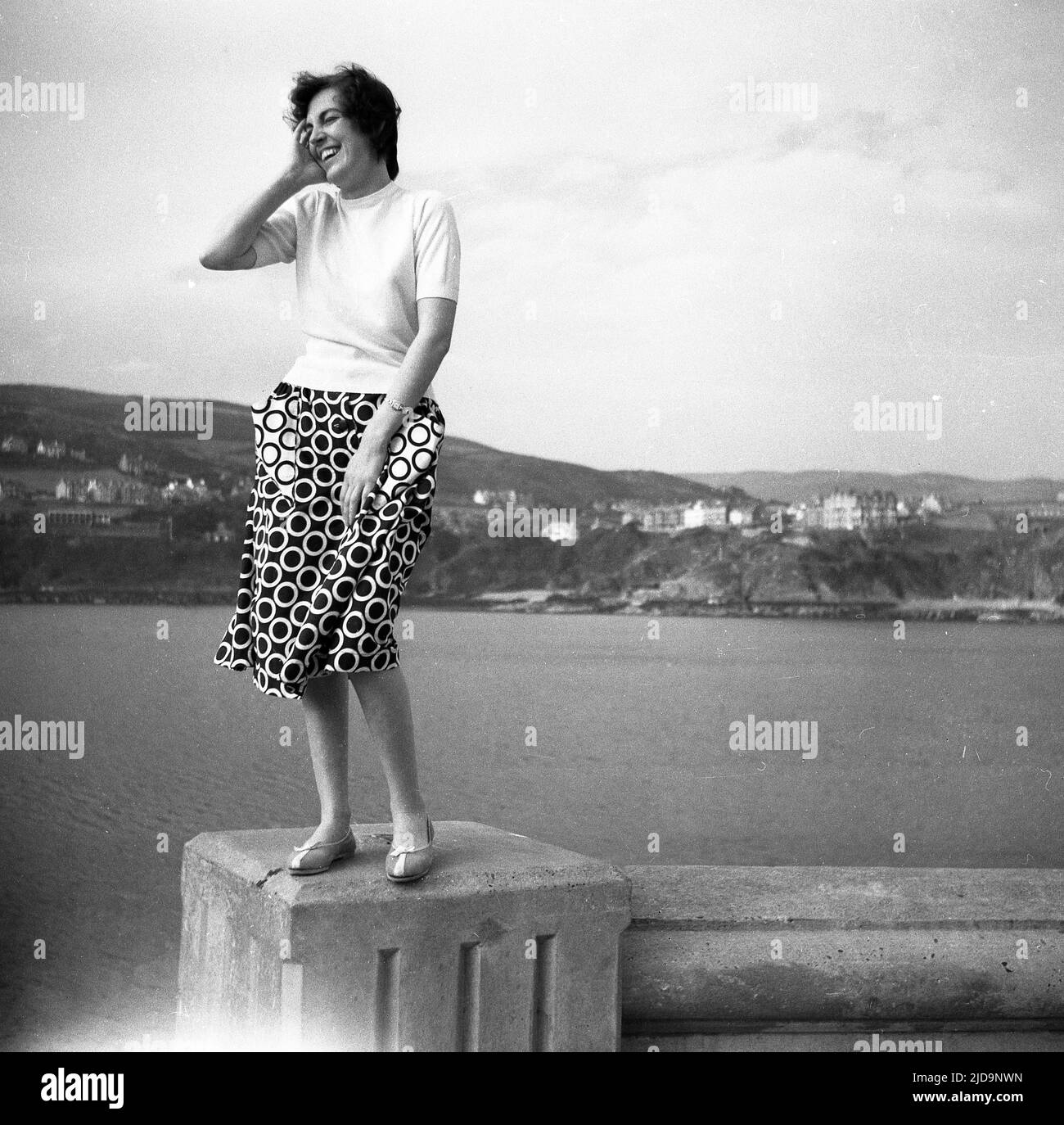 1958, historisch, eine Frau in einem gepunkteten Rock, lächelnd, während sie für ein Foto auf einem Betonposten an einem Hafen an der Küste, England, Großbritannien, steht. Stockfoto