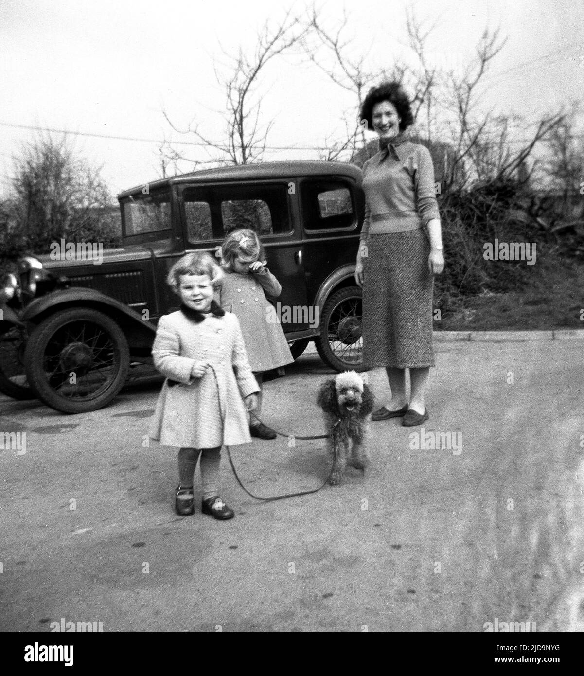 1950s, historisch, neben einem Auto der vorherigen Ära, möglicherweise einem Austin 7 oder Baby Austin, zwei junge Mädchen in Mänteln draußen mit ihrer Mutter und mit ihrem Haustier Poddle, die auf einem Doppelhund von den Mädchen geführt gehalten werden. Stockfoto