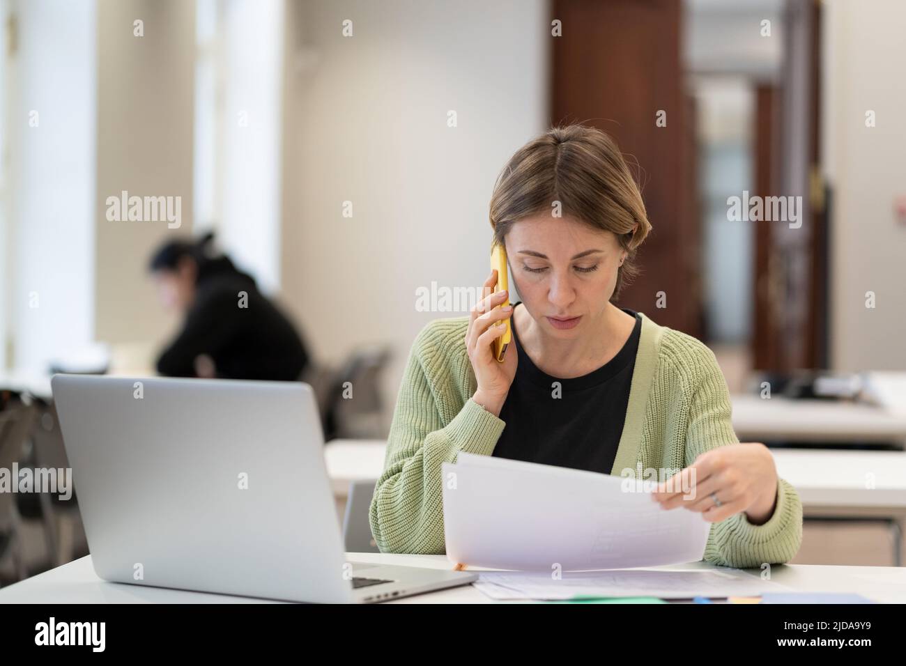 Frau mittleren Alters, die mit Dokumenten beschäftigt ist, die auf dem Mobiltelefon sprechen und auf dem Campus arbeiten. Bildung. Stockfoto