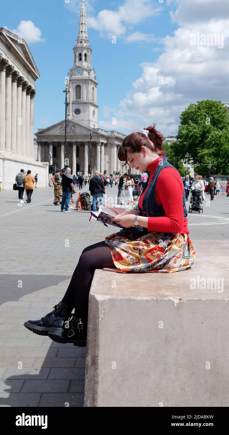 London, Greater London, England, 08 2022. Juni: Lady in farbenfroher Kleidung liest auf dem Trafalgar Square ein Buch mit St. Martins auf den Feldern dahinter. Stockfoto