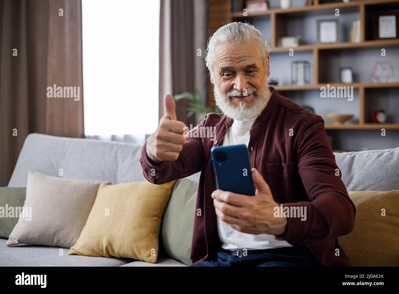 Lächelnder älterer Mann, der auf der Couch sitzt und den Daumen hochgibt, während er Computerspiele spielt, die Kamera anschaut und lächelt. Stockfoto