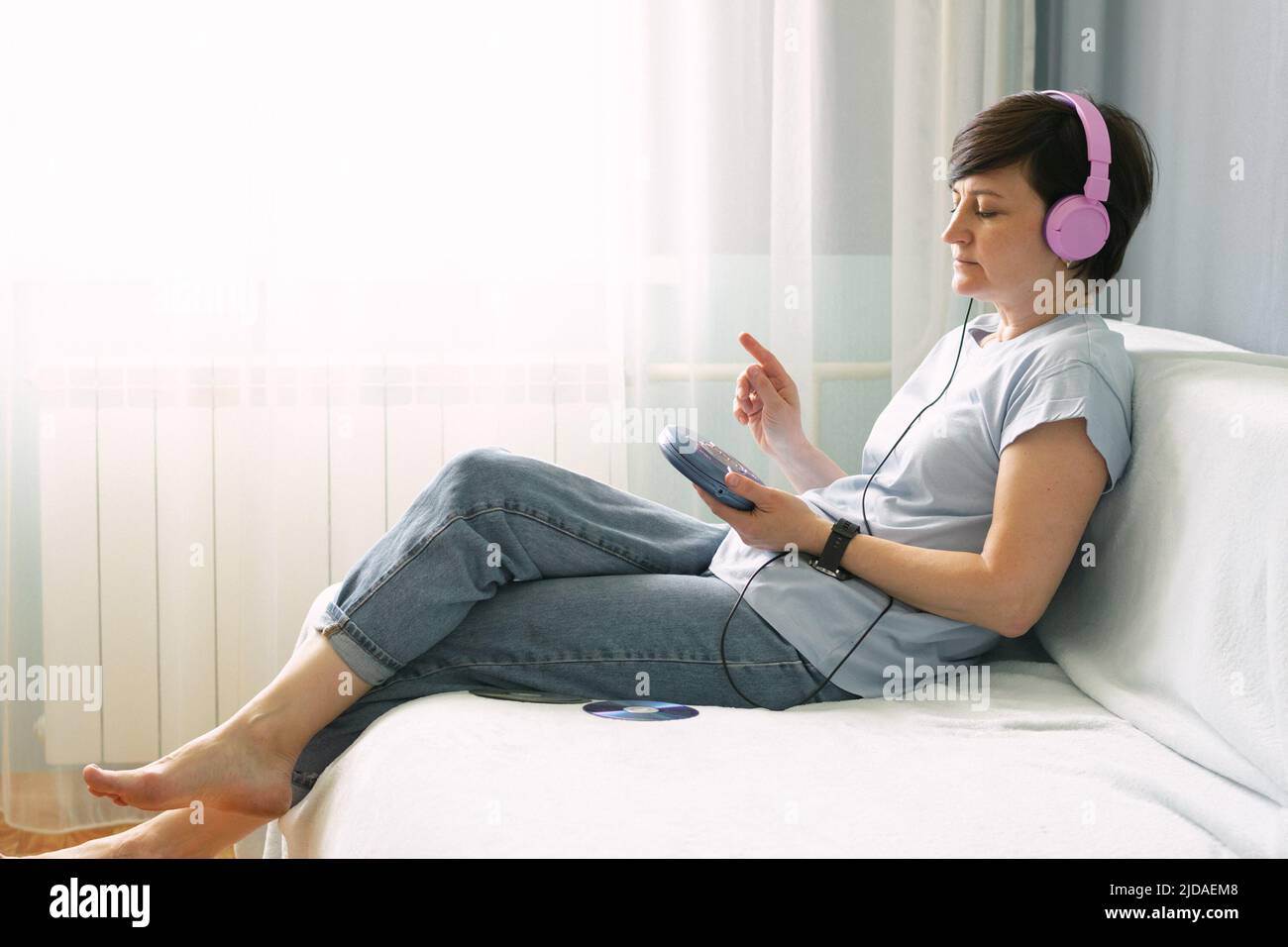 Eine Erwachsene Frau mit rosa Kopfhörern sitzt zu Hause auf dem Sofa und hört Musik auf den CD-Playern Stockfoto