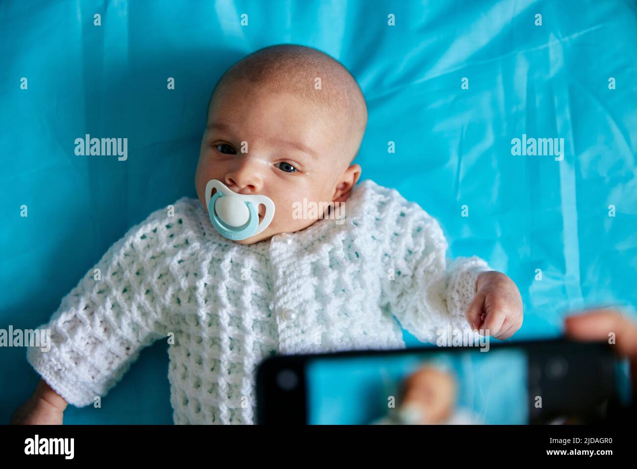 Eltern fotografieren das Baby mit Schnuller im Mund mit dem Smartphone, aufgenommen von oben Stockfoto