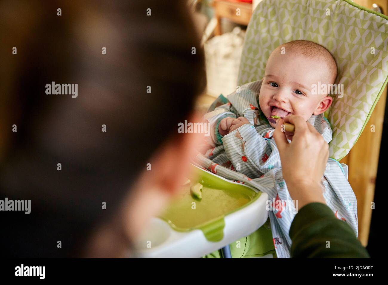 Lächelndes, sechs Monate altes Baby, das von der Mutter im Hochstuhl gefüttert wird Stockfoto