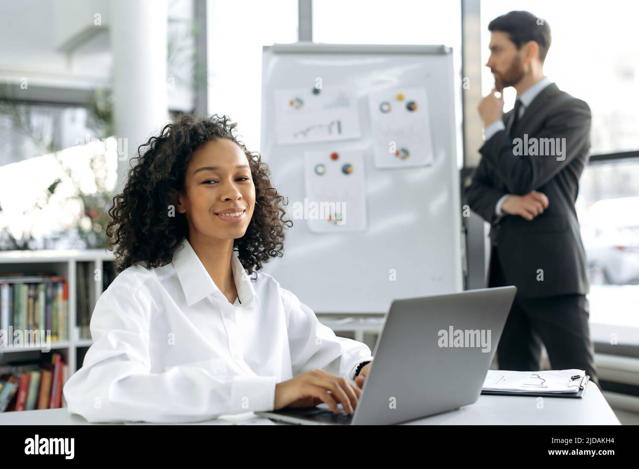 Die smarte, positive, dunkelhäutige Geschäftsfrau, Webdesignerin, sitzt an ihrem Schreibtisch vor einem Laptop. Im Hintergrund arbeitet ein männlicher Kollege mit Graphen, entwickelt eine Strategie zur Modernisierung des Workflows Stockfoto