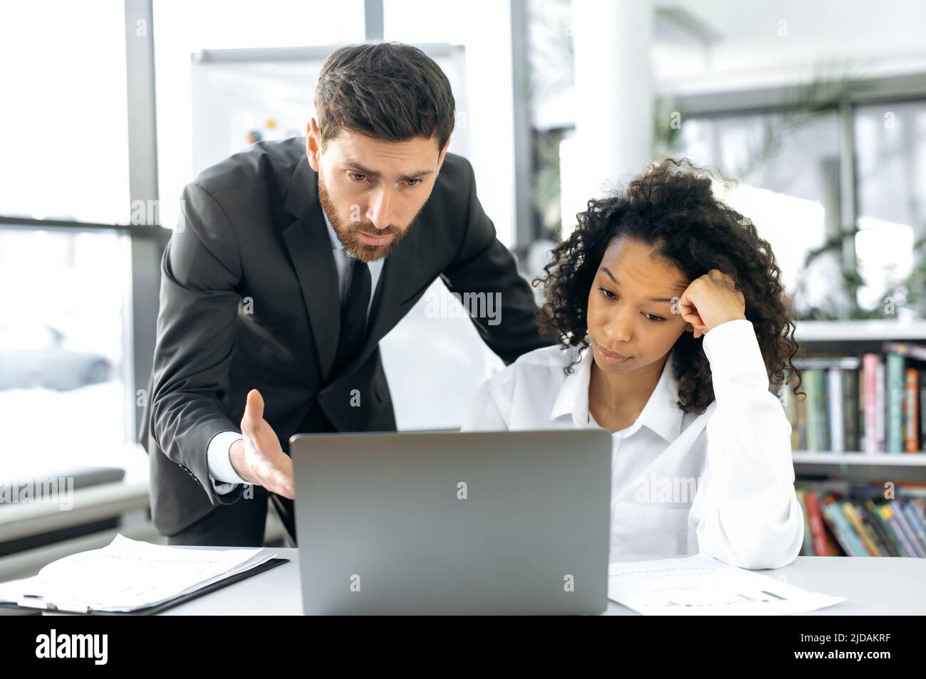 Verärgert fokussierte Kollegen aus verschiedenen Rassen, Mitarbeiter eines IT-Unternehmens, eine afroamerikanische Frau und ein kaukasischer Mann arbeiten in einem modernen Büro an einem Laptop, schauen auf den Bildschirm, testen ein neues Programm Stockfoto