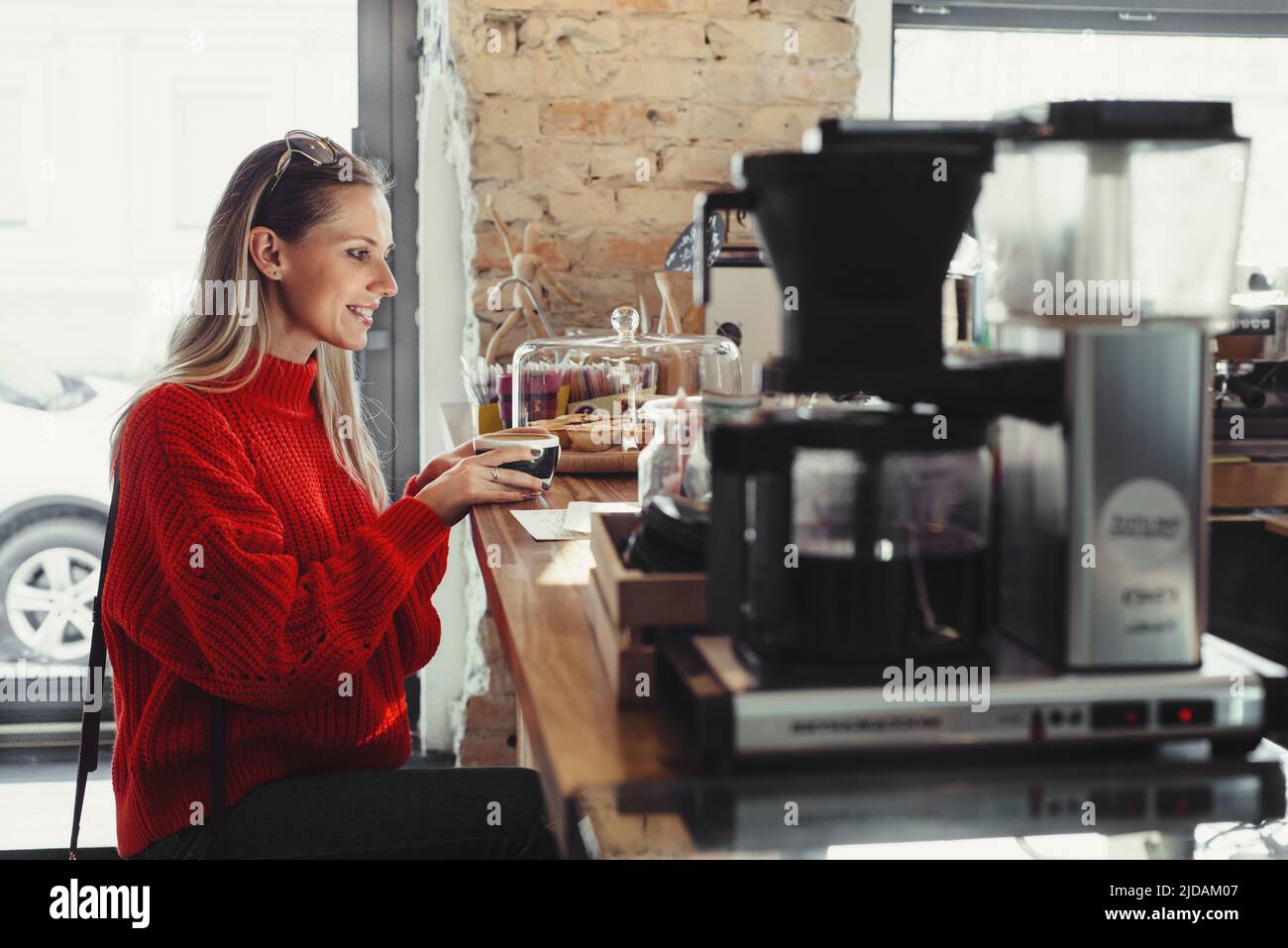 Junge, schöne Frau in rot süßem Rot, die in einem Café sitzt und einen Morgenkaffee in der Nähe der Bar trinkt Stockfoto