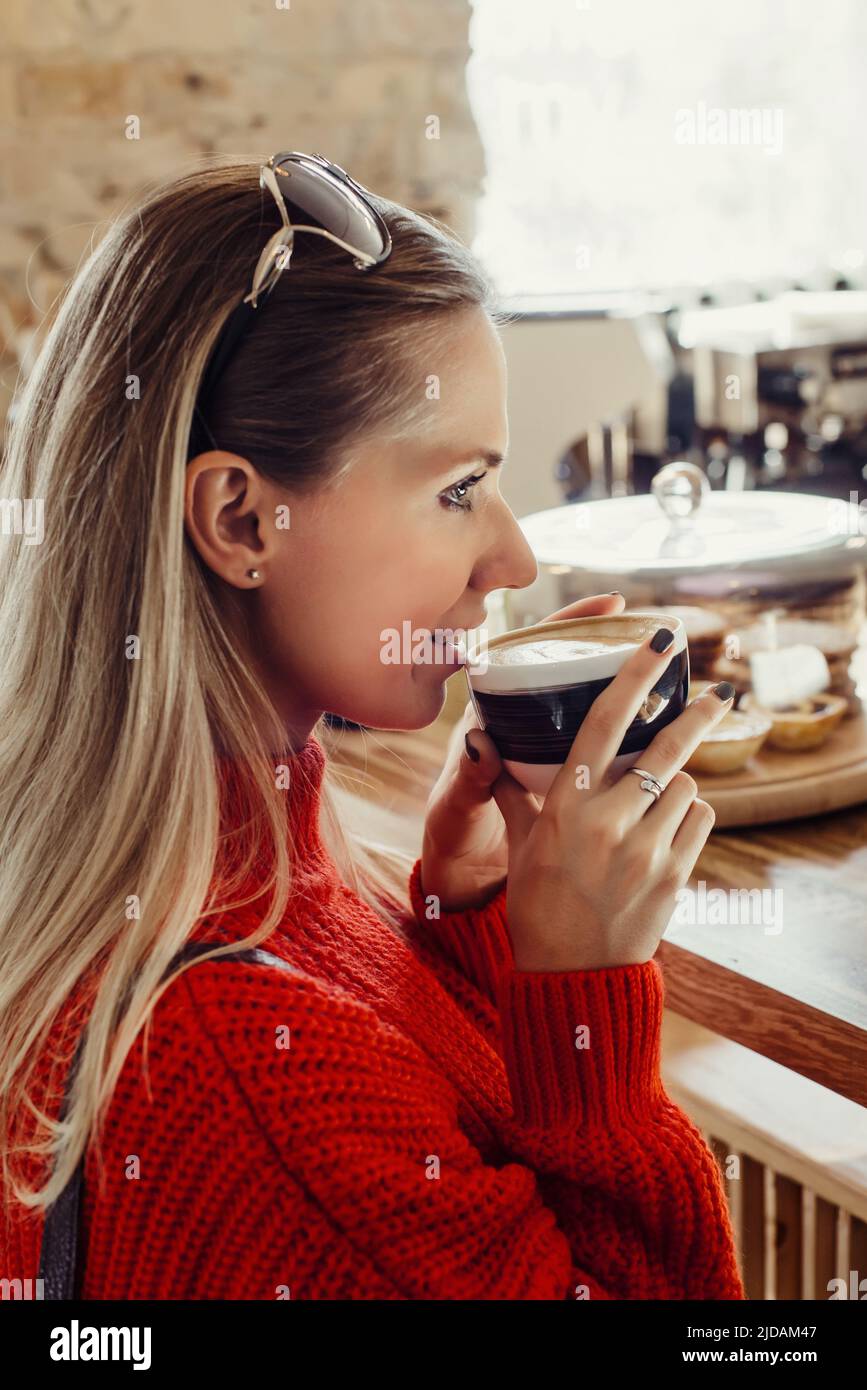 Junge, schöne Frau in rot süßem Rot, die in einem Café sitzt und einen Morgenkaffee in der Nähe der Bar trinkt Stockfoto