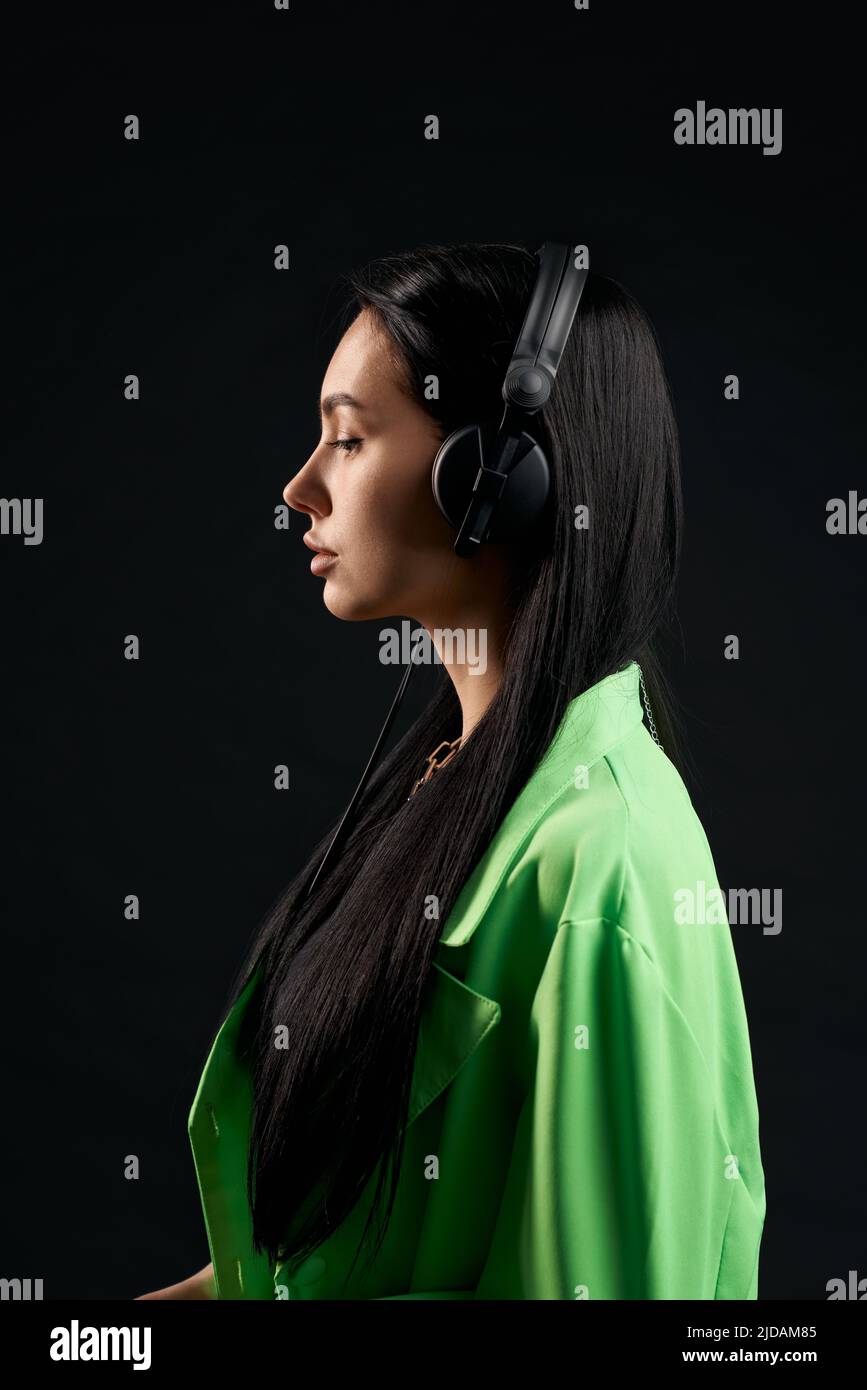 Ruhiges Mädchen in grünem Hemd, das mit Kopfhörern im Inneren Musik hört. Seitenansicht einer ernsthaften Frau mit dunklem Haar und großem über-Ohr-Headset, isoliert auf schwarzem Studiohintergrund. Konzept des Posierens. Stockfoto