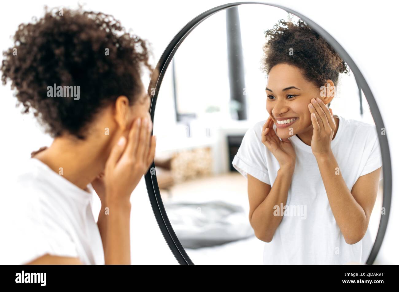 Fröhliches afroamerikanisches lockiges Mädchen in weißem T-Shirt, steht vor einem Spiegel im Wohnzimmer, lächelt fröhlich und blickt ihr Gesicht im Spiegel an, ist zufrieden mit dem Zustand ihrer Gesichtshaut Stockfoto