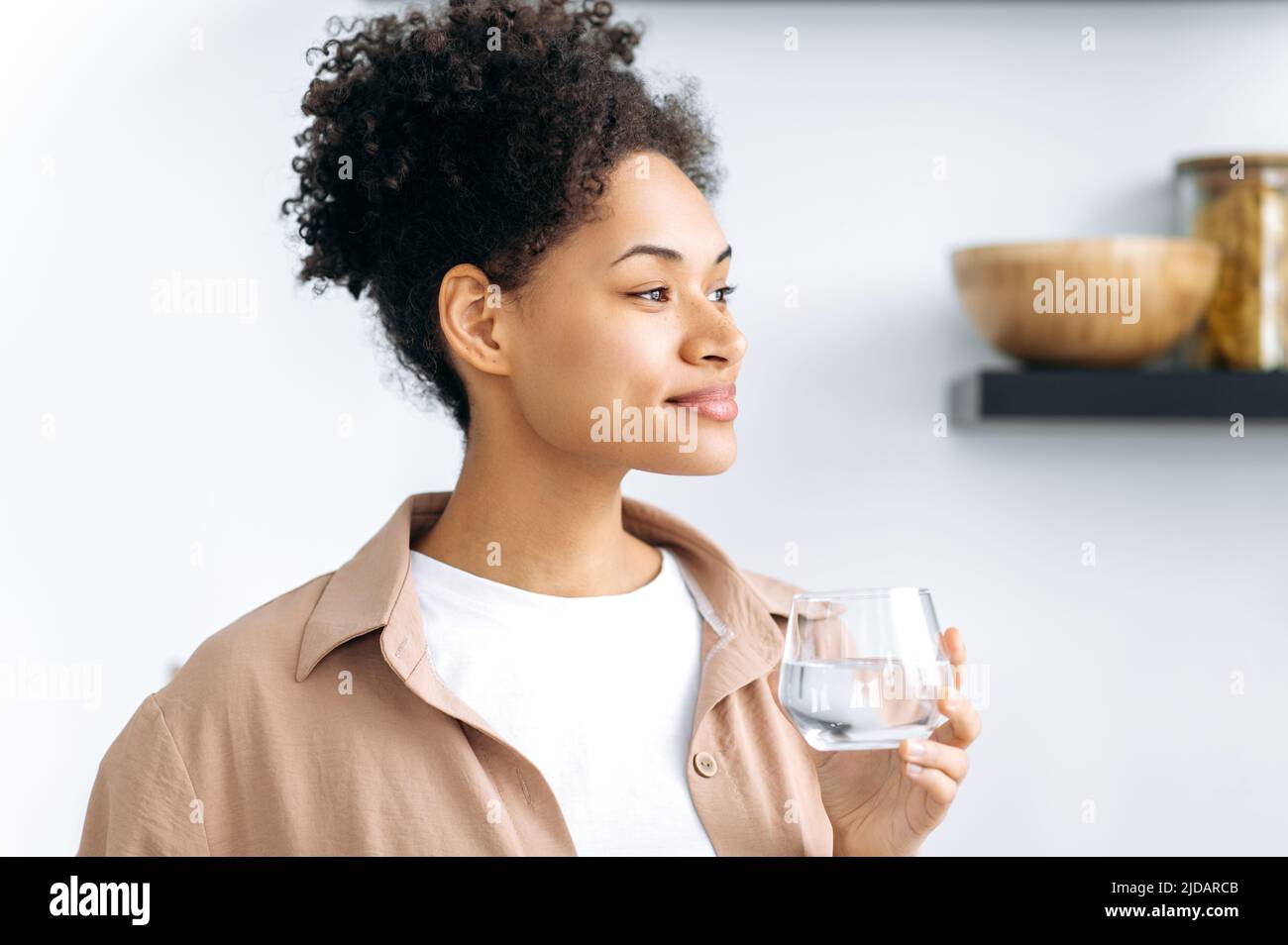 Foto eines gesunden positiven afroamerikanischen lockigen Mädchens, das ein Glas sauberes Süßwasser hält, auf die Gesundheit achten, die tägliche Körperwasserrate kontrollieren, wegschauen, träumen, lächeln Stockfoto