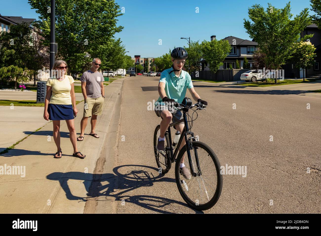 Ein junger Mann, der mit dem Fahrrad eine Wohnstraße entlang fuhr, während seine Eltern auf dem Bürgersteig standen und dabei zuschauten: Edmonton, Alberta, Kanada Stockfoto